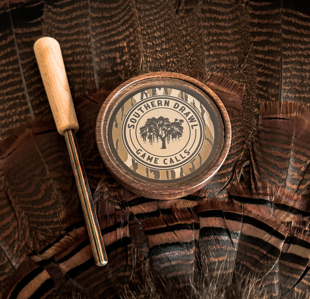 A wooden turkey pot call with a striker, branded with 'SOUTHERN DRAW GAME Calls', placed on a feathery surface.